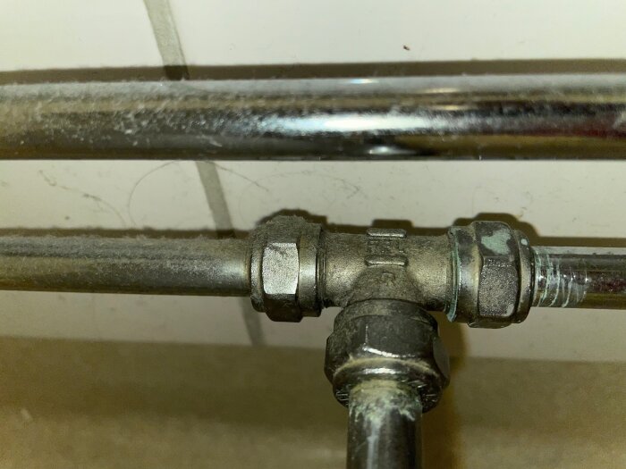 Nära bild på en t-koppling med tre metalrör anslutna, tydliga spår av vatten och förslitning syns på anslutningarna.
