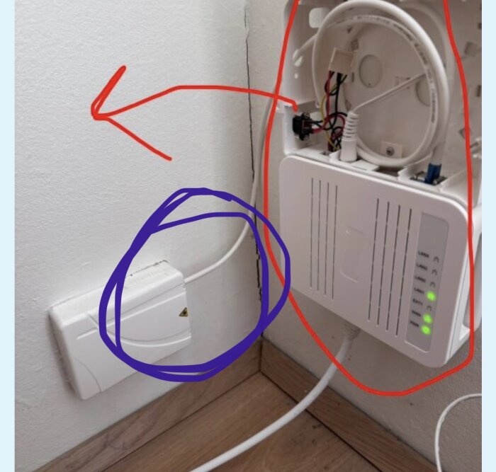 Fiberanslutning med kabel vid väggen, markerad med blå ring, nära en nätverksenhet i ID-försett skåp med öppet lock och LED-lampor.