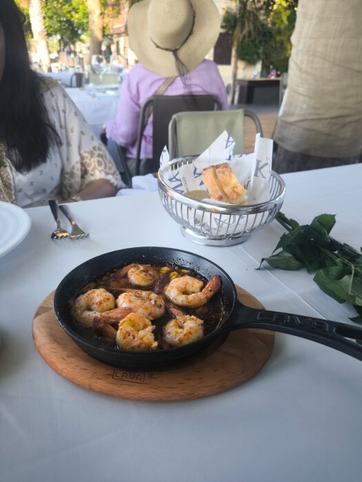 Räkor i en svart stekpanna på ett bord, med bröd i korg och två personer sittande utomhus på en restaurang.