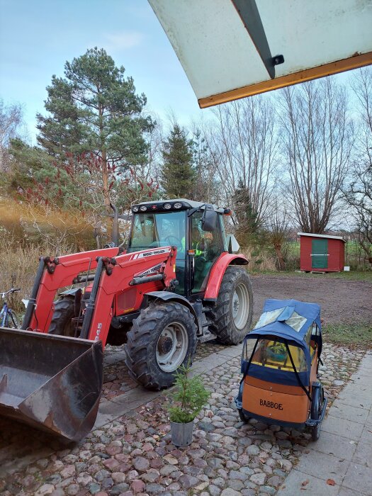 Röd Massey Ferguson traktor parkerad på stenlagd uppfart bredvid en Babboe-låda. Bakgrund med träd och en röd stuga.