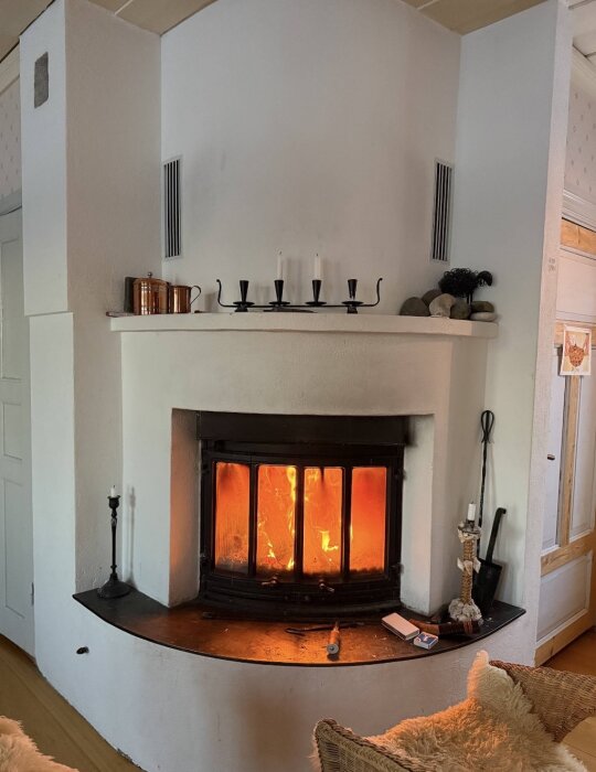Brinnande brasa i en Jøtulinsats, omgiven av en ljusstake och vedverktyg, skapar en mysig atmosfär.