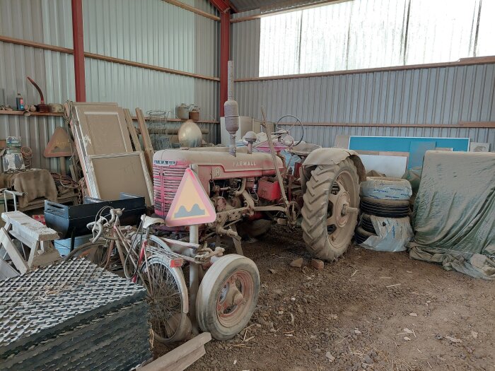 Röd Farmall traktor i en lada, omgiven av cyklar, däck och diverse föremål. Traktorn har destillatmotor utan hydraulpump.
