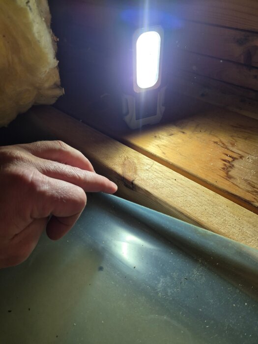 Hand pekar på plastångspärr över träbalk med isolering synlig i bakgrunden, upplyst av lampa på vinden.
