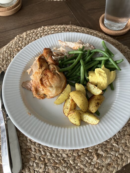 Tallrik med grillad kyckling, gröna bönor, rostad potatis och en klick sås. Glas vatten bredvid.