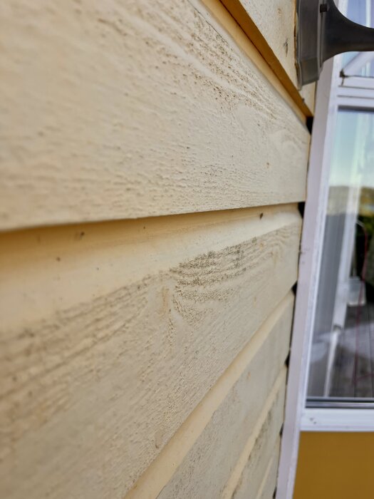 Närbild av gul fasadpanel med slät yta och lutande sektion bredvid ett fönster; detaljer av träets struktur syns tydligt.