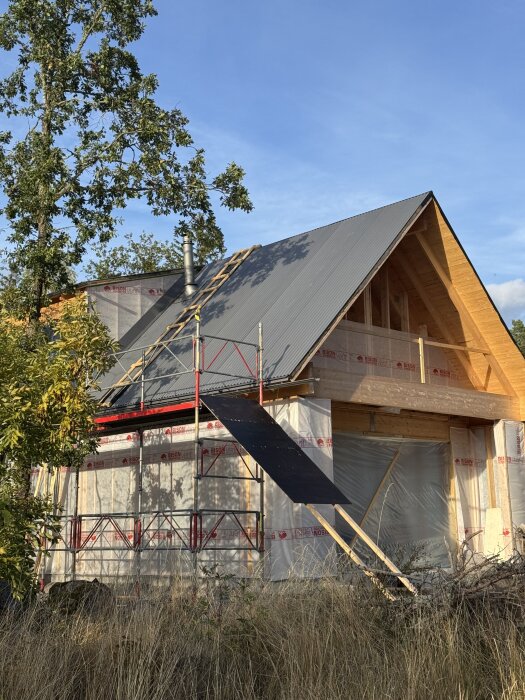 Husbygge med pågående takarbete, byggnad med ställningar runt och plåttak nästan klart. Plast täcker väggar, träd i bakgrunden.