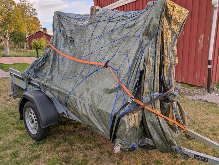 Släp med täckta glasrutor säkrade med presenning och lastnät, placerat på gräsmatta bredvid en röd byggnad.