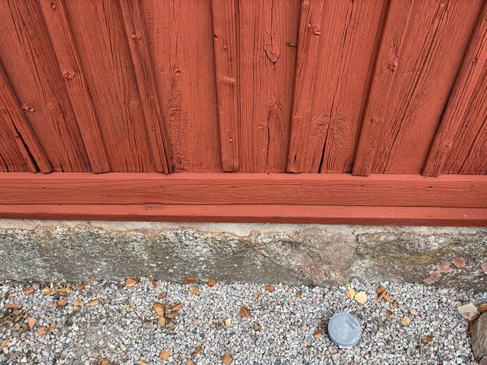 Närbild på en rötskadad röd träfasad med en tvärgående planka längst ner och en stenfot med grus framför.