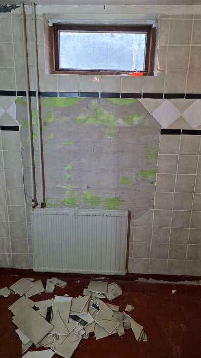 Vägg i tvättstuga med borttagna kakelplattor och synligt murbruk. Trasiga plattor ligger på golvet framför en radiator.