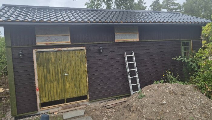 Ett nybyggt förråd/verkstad/mc-garage i trä med svart tak och gröna dörrar. Stege lutad mot väggen, omgiven av grönska och jordhög.