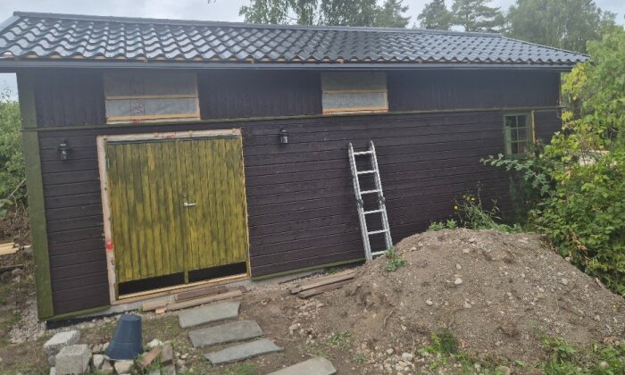 En byggnad med nyuppsatt fasad, utan foder och salningar runt dörrar och fönster. Stege lutar mot väggen, jordhög framför.