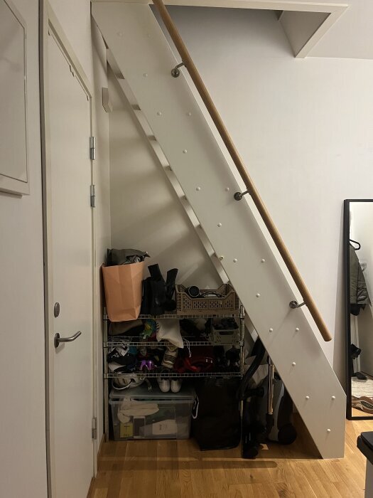 Utrymme under en vit trappa med trähandräcke, fylld med skor och saker, del av en diskussion om att bygga en platsbyggd dörr.