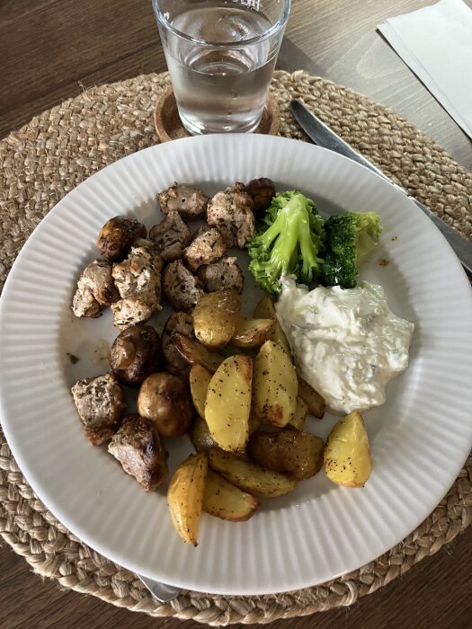 Tallrik med souvlaki, klyftpotatis, broccoli och tzatziki. Glas vatten vid sidan.