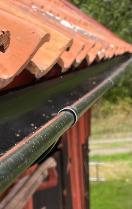 Hängränna med takpannor i bakgrunden, monterad på sidan av en lada vid ett fritidshus.