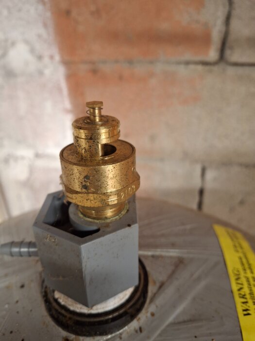Närbild på en mässingsventil på en behållare, eventuellt en hydrofor, med en grå fästkomponent mot en vägg av tegelsten.
