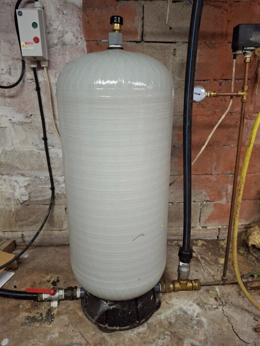 En stor grå cylinderformad enhet, förmodligen en hydrofor, står på ett cementgolv nära en vägg med ventiler och vattenrör anslutna upptill och nedtill.