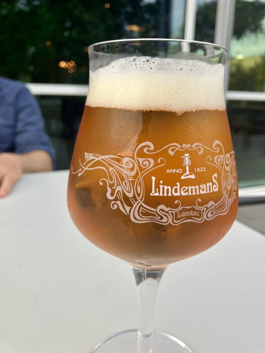 Ett glas med bärnstensfärgad Lindemans Lambic-öl, placerad på ett vitt bord utomhus med suddig bakgrund.