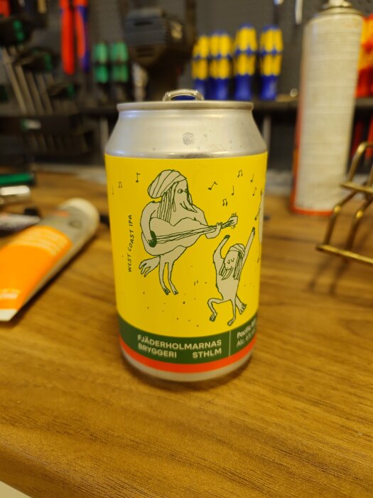 Ölburk med färgglad etikett från Fjäderholmarnas bryggeri, illustrerad med figurer. Står på en arbetsbänk i ett garage med verktyg i bakgrunden.