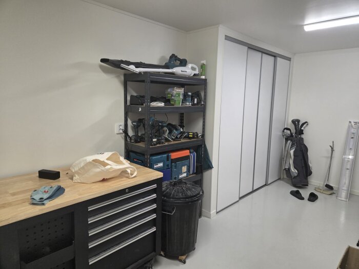 Verkstad med arbetsbänk, hylla med verktyg, soptunna och golfklubbor mot en vit vägg.