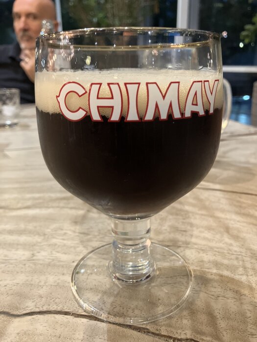 Ett stort glas Chimay-öl på ett bord, med suddig bakgrund och en man i bakgrunden.