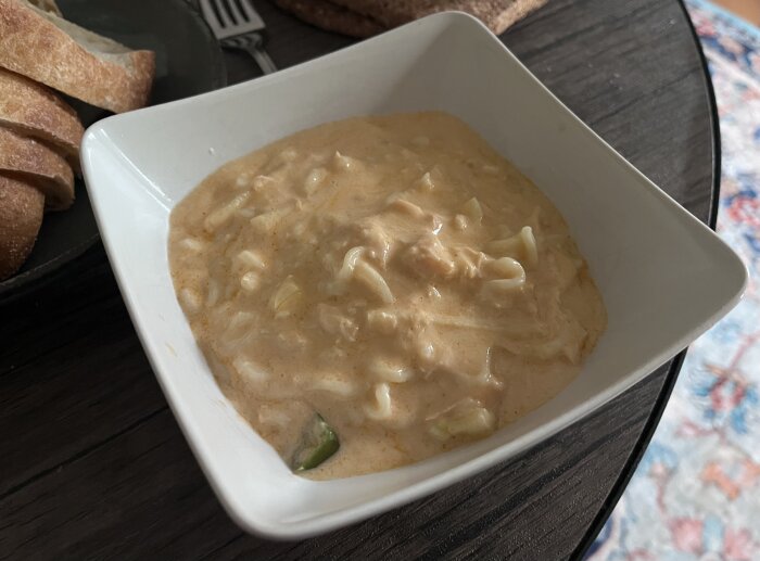 Krämig soppa i en vit skål med lax och nudlar, serverad med skivad ciabatta på ett mörkt bord.
