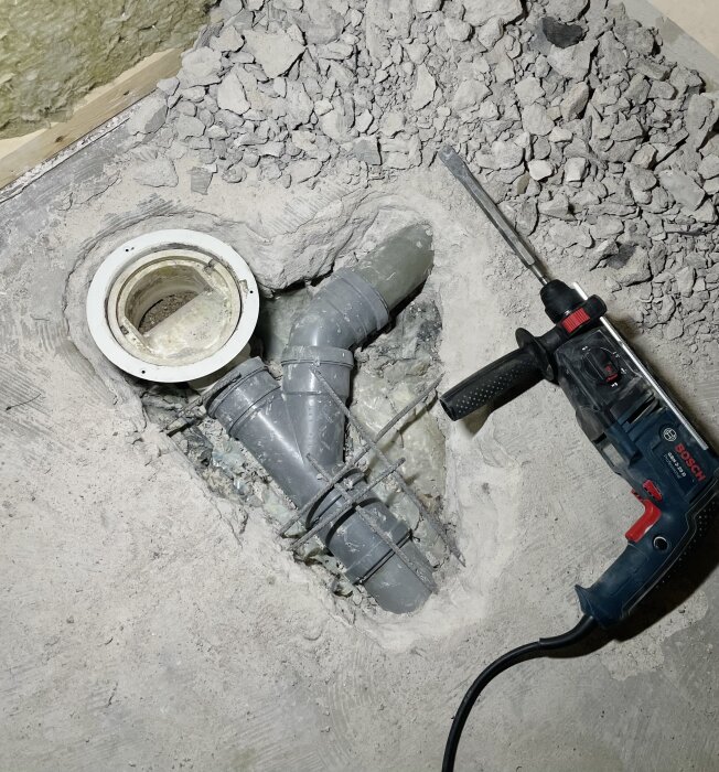 Borrmaskin bredvid öppen golvbrunn och rör, omgiven av sönderslagen betong, i ett badrum under renovering.