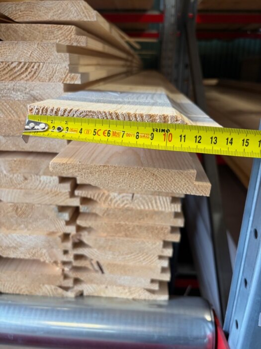 Träpaneler staplade på hylla, med en tumstock mäter ett spår som är ungefär tre centimeter långt, troligen en Norgepanel.
