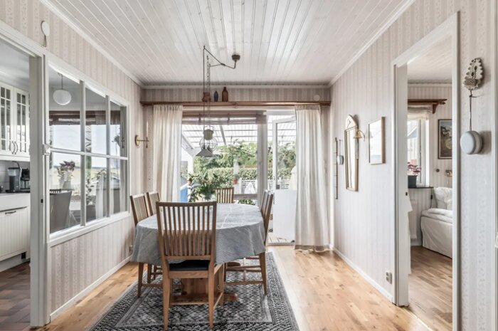 Matsal med träbord och stolar, ljusa väggar och tak, stora fönster med utsikt mot trädgården.