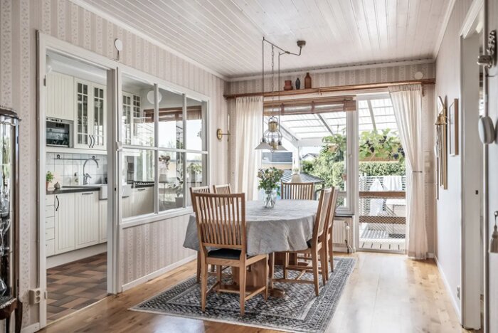 Matplats med träbord och stolar, grå duk och blommor i vas. Stort fönster och dörr mot verandan med utsikt över en trädgård. Ljus och luftig inredning.