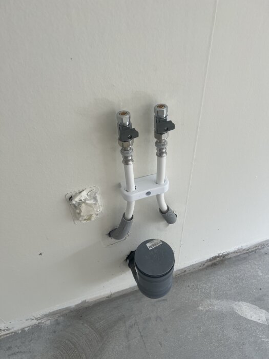 Vattenrörsinstallation på vägg med två anslutningar, gråa skyddsslangar och svart trumma nära golvet.
