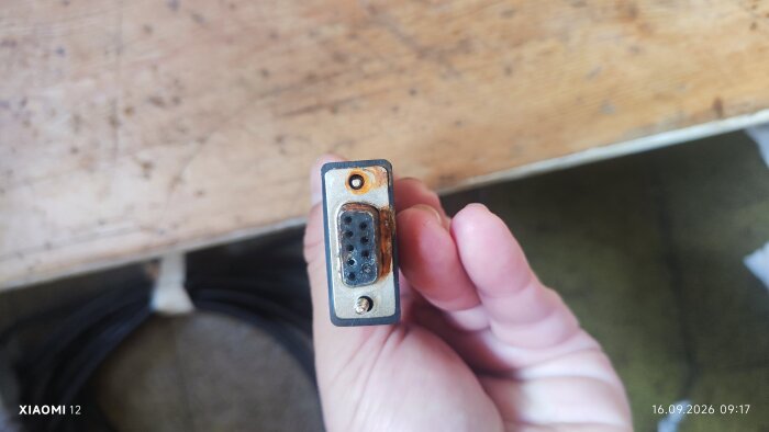 Närbild av en äldre D-sub-kontakt med rostfläckar, hålls i en hand mot en träbänk.