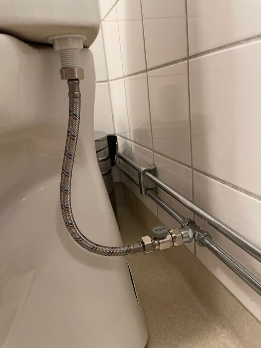 Ett VVS-system med metalliserad slang, T-koppling, bussning, kulventil och anslutning till en toalett med vita kakelväggar i bakgrunden.