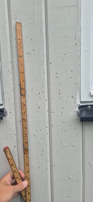 Vit träfasad med svarta små prickar och en hopfällbar mätsticka. Prickarna misstänks bero på spindlar nära garaget.