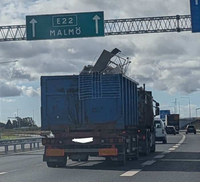 Lastbil på E22 motorvägen mot Malmö med blå container, fylld med metallskrot, under en skylt med riktningarna.