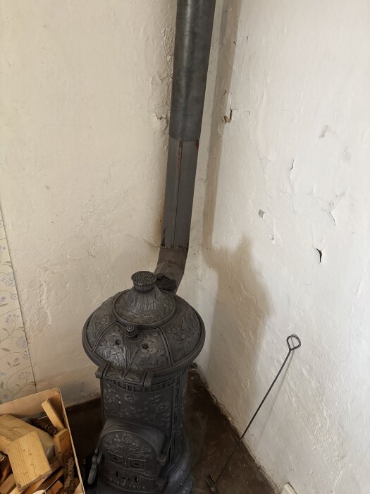 Gammal järnkamin från 1890-talet med rör nära vit, flagnande vägg; vedlåda och eldgaffel syns bredvid.