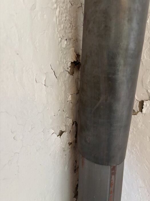 Vägg med avflagningar bredvid ett metallrör, där färg och material har lossnat, vilket antyder påverkan av värme från rökkanalen.