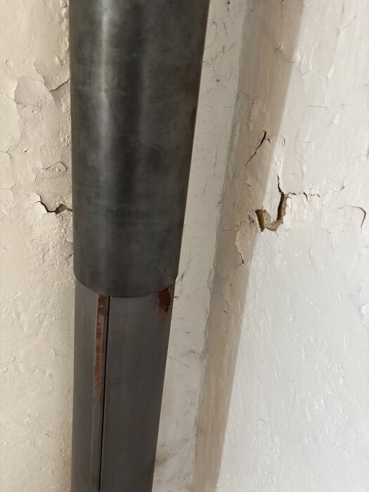 Rostigt rökrör nära en vit vägg med avflagat färglager synligt, vilket visar sprickor och underliggande skikt.