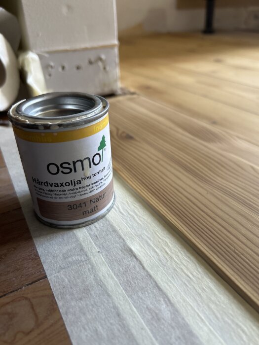 En burk Osmo hårdvaxolja 3041 Natur matt står på ett trägolv bredvid vit målartejp.