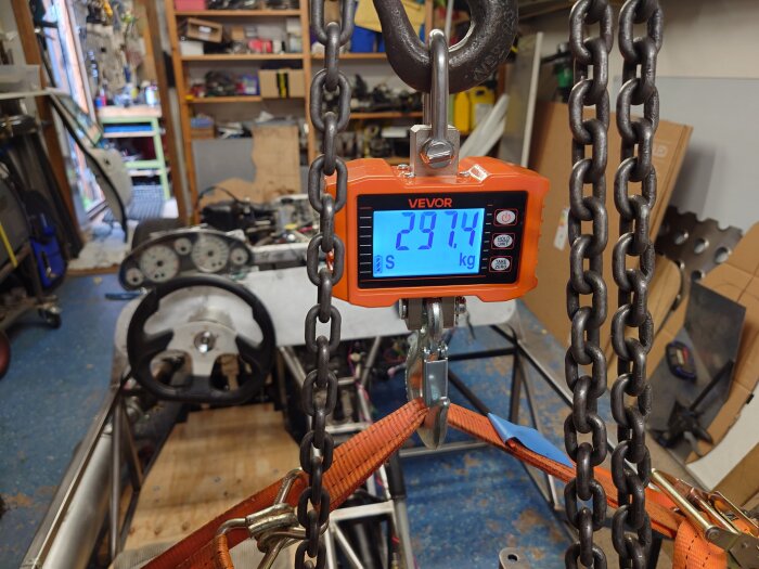 Vevor våg visar 297,4 kg i ett garage där en bils ram med ratt syns i bakgrunden. Kedjor och spännband hänger vid vågen.