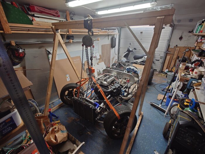 Byggprojekt i garage med motor och hjul på ram under montering, omgiven av verktyg och material. En lyft används för att hålla mekanismen.