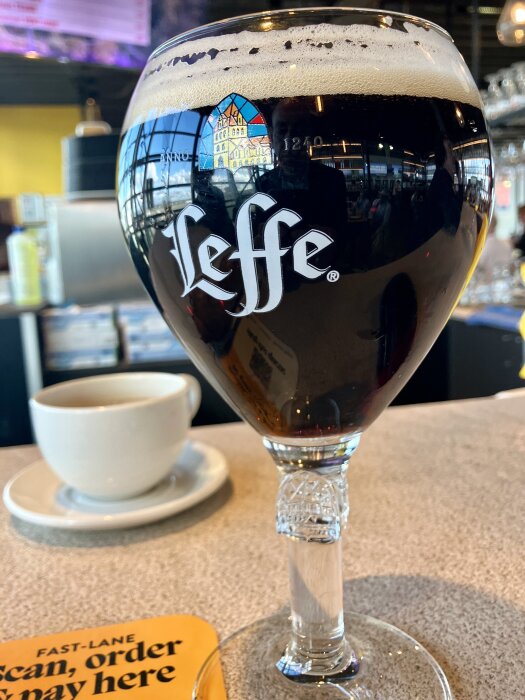 Ett glas mörk Leffe-öl med skum på en bardisk, en kaffekopp i bakgrunden och en skylt med texten "Fast-lane, scan, order & pay here".