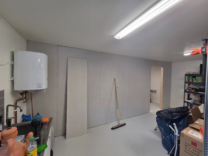 Ett garage används som föråd, med verktyg, kartonger, en soppåse, en varmvattenberedare och hyllor med förvarade föremål.