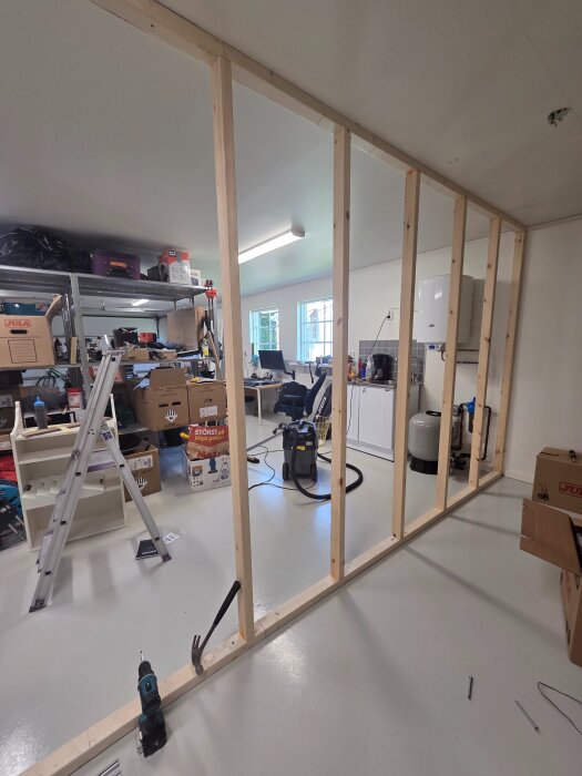Halvfärdig trävägg i garage med byggmaterial och redskap; kartonger och en stege i bakgrunden.