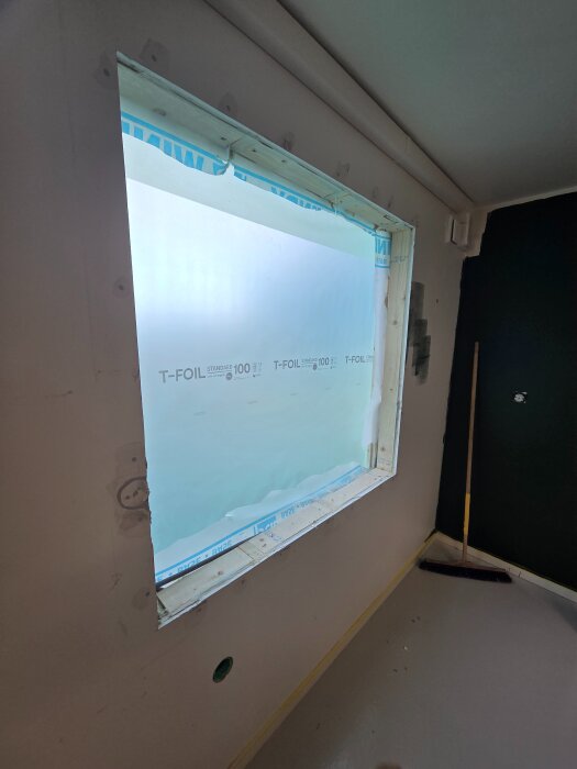 Öppning för nytt fönster i en vägg, delvis installerad med skyddsplast och ram, nära färdigställd inomhusrenovering.