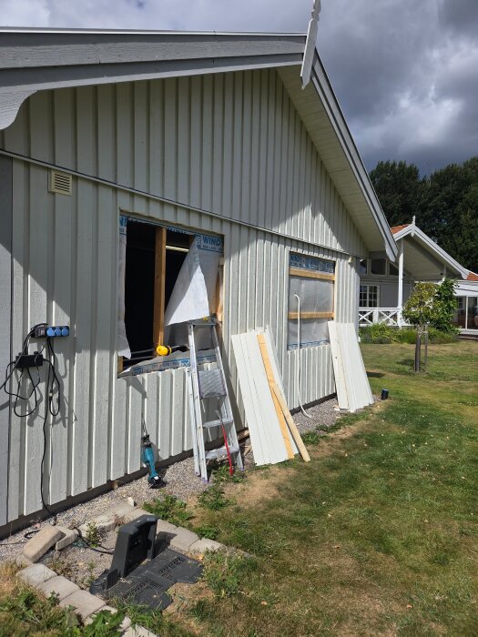 Husfasad med ett fönster under installation, stege lutad mot väggen, verktyg och brädor på marken.