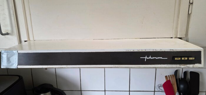 Gammal köksfläkt utan filter installerad under ett skåp, märkt med "Futurum". Tydliga kontrollknappar synliga.
