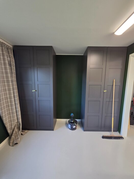 Två mörka garderobsdörrar står på varsin sida av en mörkgrön vägg, med ett fönster med rutiga gardiner till vänster. På golvet syns en sopborste och en borrmaskin.