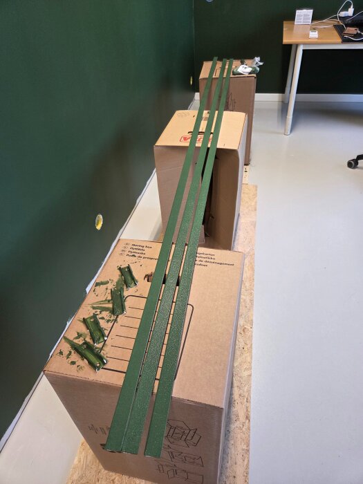 Gröna lister ligger på två flyttlådor framför en mörkgrön vägg i ett rum inrett med ett bord och ett hörn av en stol synlig.
