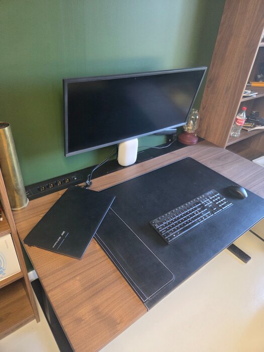 Ikea-skrivbord med en datorskärm, tangentbord och mus, mot en grön vägg med kabelhantering under skärmen.
