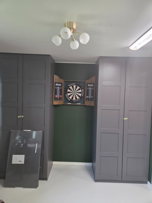 Gröna väggar med mörkgrå garderober och en darts tavla. Taket har en vit lampa, och golvet är epoximålat vitt.